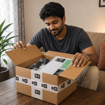 A man sitting on a couch in a living room opens a brown cardboard box sealed with white BO tape, revealing packaged items inside.